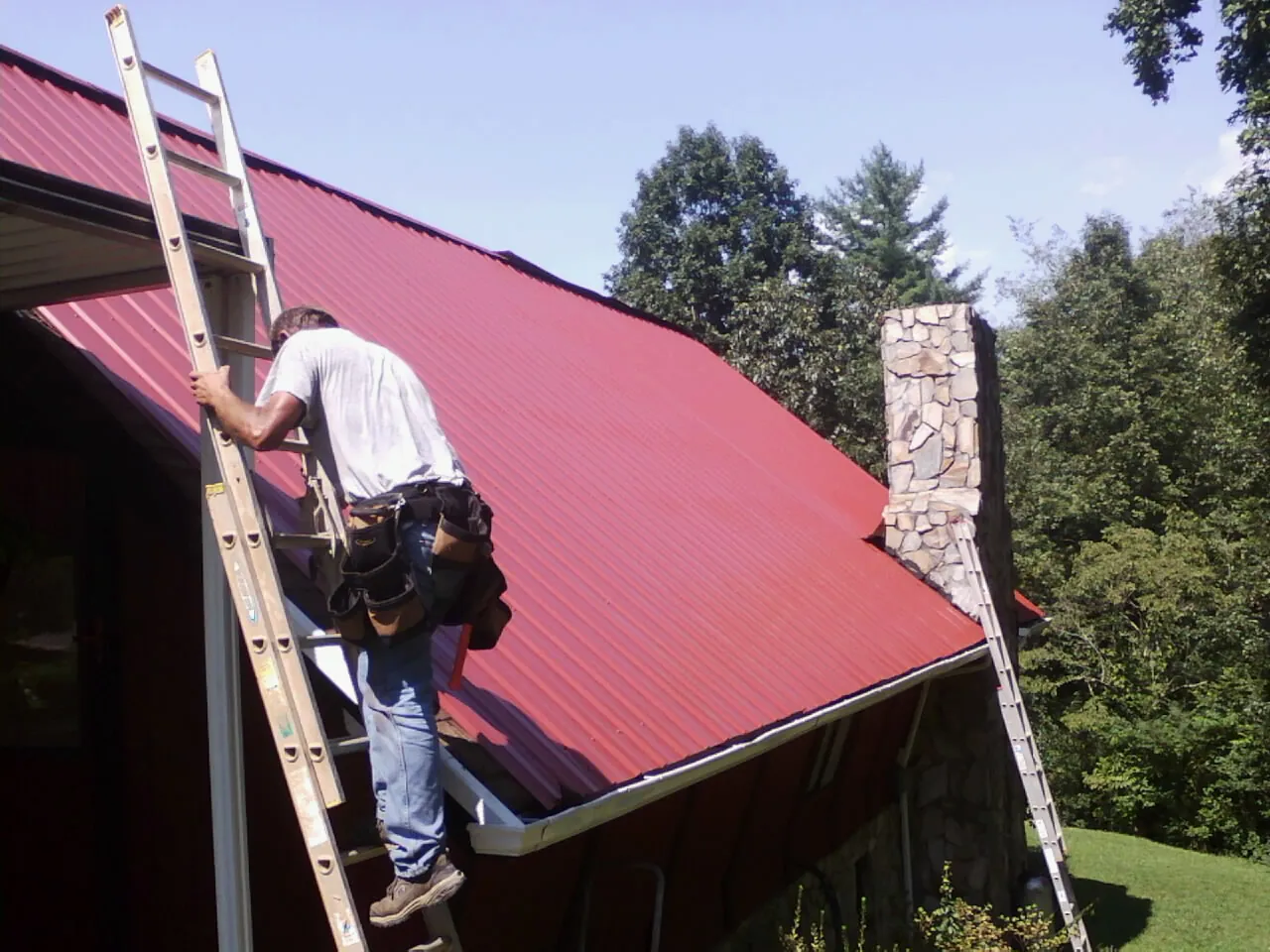 Roofer on a ladder beside a red metal roof and stone chimney — Soffit & Fascia Repair by Roofing Brothers in Smyrna